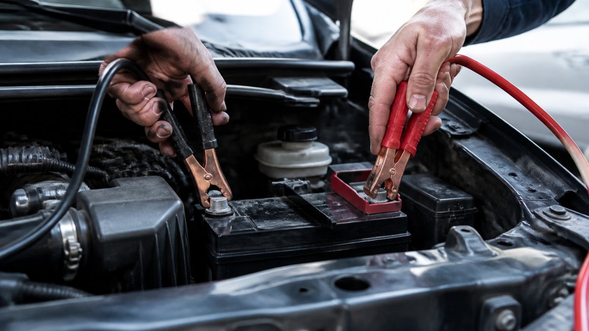 Close-up of jumper cables being connected to battery terminals, showing red cable on positive terminal and black cable on negative terminal with proper hand positioning
